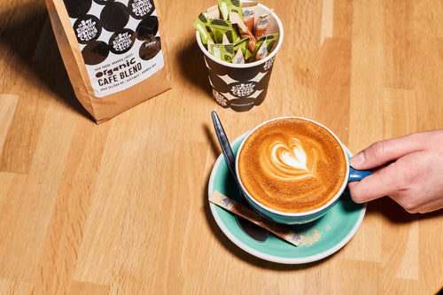 A flat white with a heart in the milk beside a bag of Bay Espresso organic Cafe Blend
